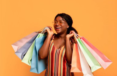 Memnun bir müşteri. Yazın neşeli genç siyahi kadın elinde parlak alışveriş çantaları tutarak poz veriyor ve yan bakıyor, turuncu stüdyo arka planında yaptığı alışverişlerden memnun.
