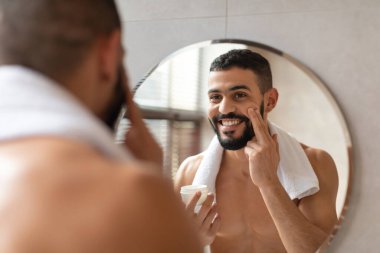 Modern bir banyoda yakışıklı bir adam yüzüne nemlendirici krem sürerken yansımasına gülümser. Kendine güveni tam bir şekilde duruyor. Cildine özen gösteriyor..