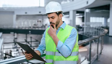 Güvenlik teçhizatı alanında çalışan bir inşaat işçisi, binanın bitmemiş büyük bir yerinde dururken planları gözden geçirir. İşçi bilgiyi incelerken düşünceli görünüyor..