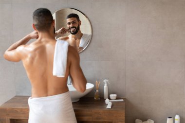 Mutlu genç bir Arap, parlak ve modern bir banyoda dişlerini fırçalıyor. Havluya sarılmış, aynaya bakıyor. Yer temiz ve iyi organize edilmiş, kişisel hijyeni teşvik ediyor..