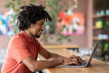 Gülümseyen kıvırcık, çekici siyah erkek bilgisayarda yazı yazıyor, kafenin içindeki sosyal ağlarda sohbet ediyor. Bağımsız yüklenici için uzak işyeri, serbest çalışma, modern iş ve teknoloji