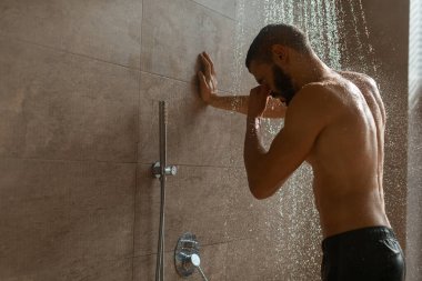 Genç sakallı bir adam şık bir banyoda sıcak suyun altında duruyor. Vücudunu yıkıyor ve kendine bakıcılık yapıyor. Atmosfer sakin ve gençleştirici, günlük hijyen rutinleri için mükemmel..