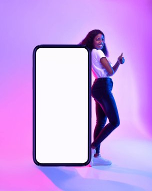 Güzel bir Afro-Amerikalı kadın başparmağını kaldırırken dev bir cep telefonuna yaslanıyor. Neon ışıkları canlı bir atmosfer yaratarak yeni bir mobil uygulama veya web sitesi oluşturur..