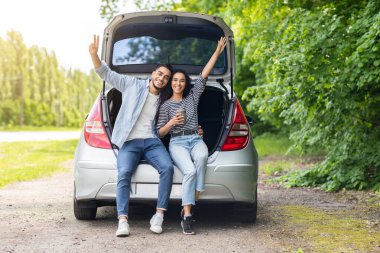Tasasız çok ırklı çift, araba gezisi sırasında kırsalda mola veriyorlar, açık bagajda oturuyorlar, kahve içiyorlar, kucaklaşıyorlar ve neşeyle kameraya gülümsüyorlar, el kaldırıyorlar, boş alan.