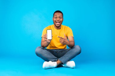 Excited Black Guy Showing Smartphone Pointing Finger At Blank Cellphone Screen Advertising And Recommending New Mobile Application Sitting Over Blue Studio Background. Mockup