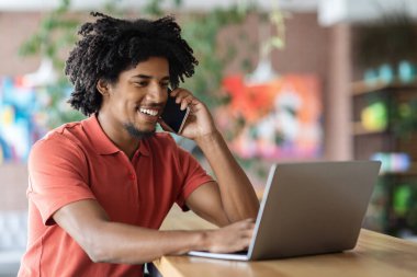 Cep telefonuyla konuşan, kafede dizüstü bilgisayar kullanan, bilgisayarla uzaktan çalışan ve cep telefonu sohbetinin keyfini çıkaran gülümseyen Afrikalı Amerikalı adam,