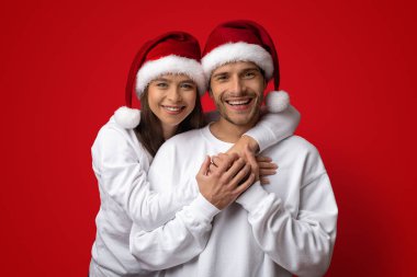 Mutlu bir çift, Noel Baba şapkaları giyerken kucaklaşıp gülümsüyor. Canlı kırmızı bir arkaplanda toplanırlar, tatilin neşeli ruhunu yakalarlar..