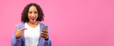 Kıvırcık saçlı, gülümseyen ve akıllı telefonu işaret eden bir genç. Arka plan pembe. Açık renk bir gömlek ve mor bir hırka giyiyor. Heyecan ya da neşe gösteriyor..