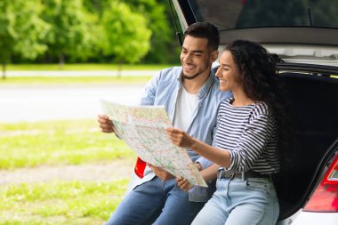 Orta Doğulu çiftin yol haritasını izlemesi, mola vermeleri, arabayla birlikte seyahat etmeleri, fotokopi alanı olan panorama. Gülümseyen Arap adam ve güzel adam açık bir bagajda oturmuş, yeri kontrol ediyorlar.