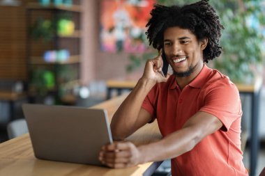 İyi ki Afro-Amerikan kıvırcık erkeği telefonla arayıp kafenin içindeki masada dizüstü bilgisayara bakıyor. Uzaktan çalışma ve iş yerindeki modern çalışma teknolojisi ve dijital göçebe ile iletişim kurma