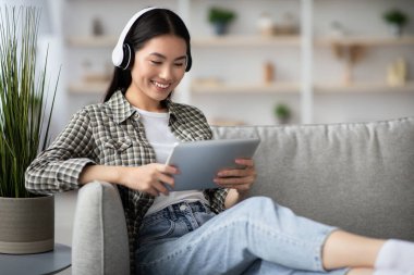 Güzel Asyalı kadın film izliyor, evdeki kanepede dinlenirken kablosuz kulaklık ve ped kullanıyor. Mutlu genç kadın dijital tablette eğlenceli yeni mobil uygulamalarla eğleniyor