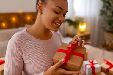 Noel ve yeni yıl kutlaması. Mutlu Afro-Amerikan bayanı Noel hediyesini açıyor ve gülümsüyor, yatakta oturuyor ve kırmızı kurdeleli hediye paketini açıyor.