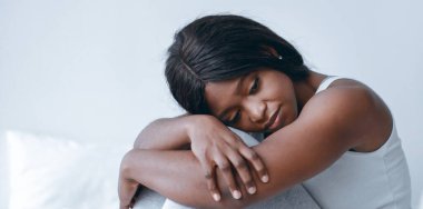 Genç bir kadın başını eğip kollarını kavuşturarak oturur. Yumuşak bir ışığın geldiği parlak bir odada dinlenirken düşünceli ve düşünceli görünüyor..