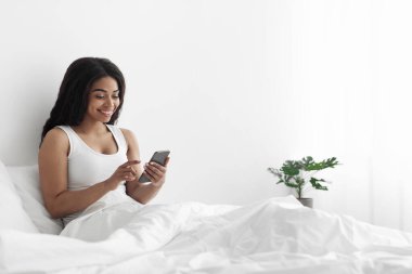 Mutlu Afro-Amerikan kadın blogcu akıllı telefonuyla yazı yazıyor, yatakta oturuyor, boş yer var. Sosyal ağlar, internette sörf, haber ve hafta sonları evde sohbet.