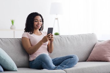 Ev içi rahat, modern telefon kullanan, sosyal medyada cihaza mesaj yazan bir kadın. Çevrimiçi arayan mutlu genç siyahi bayan oturma odasının iç tarafında web kamerası olan sanal toplantı kumandasına sahip.