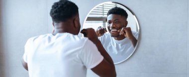 Banyoda yuvarlak aynanın önünde duran bir adam. O, yansımasına bakarken dişlerini fırçalıyor. Oda aydınlık ve modern bir tasarımı var..