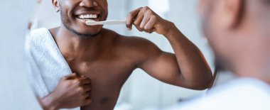 Bir adam aynanın önünde durup dişlerini fırçalıyor. O bir havluya sarılı. Banyodaki ışık parlak. Lavaboda su görülebilir..