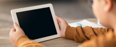 Bir çocuk masada oturuyor, elinde bir tablet var ve boş ekrana bakıyor. Masada birkaç kağıt ve bir kalem var. İçerisi yumuşak ışıklandırmayla dolu..