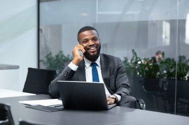 Takım elbiseli bir adam modern bir ofiste masa başında oturur. Telefonda konuşurken gülümsüyor. Önünde bir dizüstü bilgisayar ve belgeler var. Bitkiler arka planda görülebilir.
