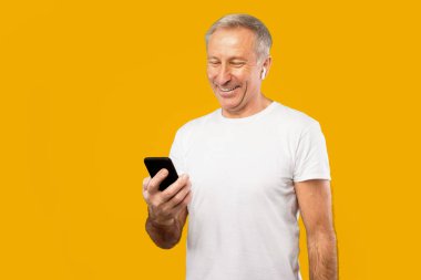 Yaşlı bir adam sarı bir arka planın önünde duruyor. O, beyaz bir gömlek ve kulaklık giyiyor. Akıllı telefonunu kontrol ederken mutlu görünüyor..