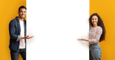 Gülümseyen Orta Doğulu çift boş el ilanı panosuna işaret ediyor, genç Arap erkek ve kadın mesajınız için kopya alanı gösteriyorlar, sarı arka planda duruyorlar, Panorama