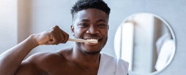 Banyoda elinde diş fırçası olan bir adam var. O, dişlerini fırçalarken gülümsüyor. Oda arka planda yuvarlak bir ayna ile aydınlık..