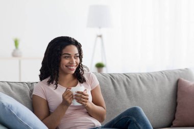 Hafta sonlarının, dinlenmenin ve kahve molasının tadını çıkarın. Boş zamanlarınızda leziz içeceklerle evde dinlenin. Gülümseyen, genç, çekici, Afro-Amerikan kadın. Oturma odasının içinde, boş bir alanda bir fincan çayla.