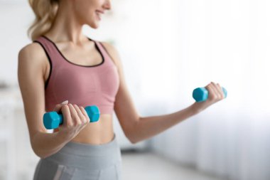 Kapsamlı 19 kilitlenme, dayanıklılık eğitimi, evde kaslara uygun. Bin yıllık neşeli sarışın kadın spor giyim kuşağında, minimalist oturma odasında halterlerle egzersiz yapıyor. İç kısımları kesilmiş, kopyalama alanı var.