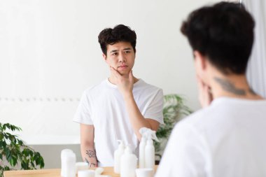 Belki tıraş olurum. Sabah prosedürleri ve erkek bakımı. Beyaz tişörtlü ciddi bir Asyalı adam. Yüzüne sürtünüyor, sakallarını kontrol ediyor ve aynadaki yansımasına bakıyor. Banyo içi boş.