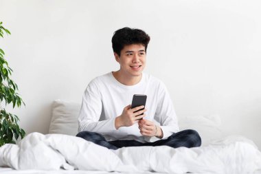 Sosyal medyada akıllı telefon ağı. Yeni mobil uygulama reklamı ve Covid 'de sosyal mesafeler. Pijamalı gülümseyen Asyalı adam akıllı telefonu tutuyor ve yatak odasındaki boş alana bakıyor.