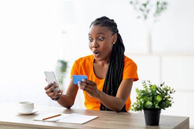 Kredi kartlı duygusal siyahi kadın şok içinde akıllı telefona bakıyor, inanılmaz bir online satış buluyor, evden alışveriş yapıyor. Şaşırmış Afrikalı Amerikalı kadın internetten mal sipariş ediyor.
