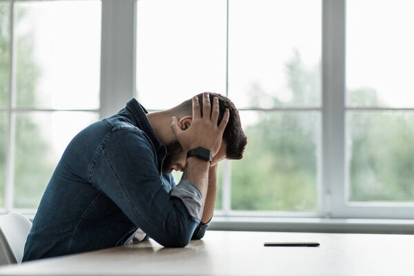 Self-isolation, depression at home, psychological problems and mental troubles. Sad millennial guy hold head sitting at table with smartphone, waiting for message on big window background, copy space