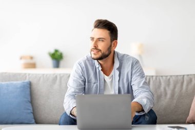 Milenyum serbest çalışanı laptopta çalışıyor ve bir kenara bakıyor, başarılı iş stratejisini düşünüyor ya da tatilin hayalini kuruyor, evdeki iç kısımda kanepede oturuyor, boş alan arıyor.