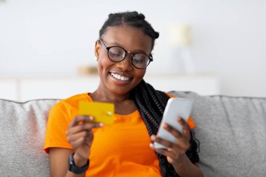 Çevrimiçi alışveriş. Kredi kartı ve akıllı telefonlu neşeli Afrikalı kadın evdeki koltukta oturuyor, uzaktan ödeme yapıyor, web mağazasından bir şeyler sipariş ediyor, müşteri hizmetlerinden memnun.