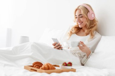 Binlerce yıllık güzel Avrupalı kadın bornozlu ve kulaklıklı. Haberler okuyor, sohbet ediyor, video izliyor ve yatak odasında yatakta kahvaltı yapıyor. Müzik, sağlık ve sağlık hizmetlerini dinleyin.