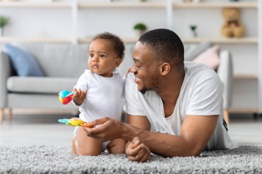 Mutlu Afrika kökenli Amerikalı baba, evde küçük bir çocukla oynuyor, küçük siyah babayla ilgileniyor, küçük bebek oğluyla oturma odasında vakit geçiriyor, oyuncakları kullanıyor ve yakınlaşıyor.