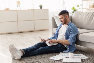 Serbest İşçi Günü 'nde. Gülümseyen milenyum çalışanı öğrenci evde çalışıyor oturma odasındaki halının üzerinde oturuyor. Elektronik evrak işleriyle uğraşan erkek bilgisayarı kullanıyor.