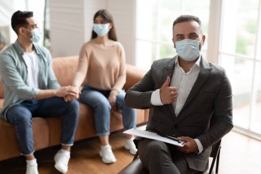 Ofisteki genç çiftle başarılı bir terapiden sonra tıbbi maske takan profesyonel erkek psikolog işaret veriyor. Eşler klinikte evlilik danışmanıyla el ele tutuşuyorlar.