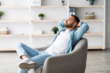 Neşeli genç adam rahat koltukta dinleniyor, gözleri kapalı rahatlıyor, evde başının arkasında el ele tutuşuyor. Pozitif beyaz adam hafta sonunu oturma odasında dinlenerek geçiriyor.