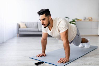 Güç egzersizi konsepti. Genç bir Arap 'ın evde şınav çekmesi ya da kalas çekmesi, akıllı telefondan video izlemesi, boş alan. Atletik Doğulu bir adam covid-19 tecridi sırasında aile içi eğitim alıyor.