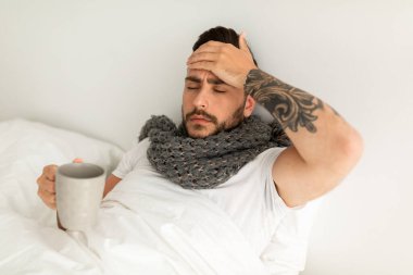 Kötü hissediyorum. Ateşli hasta bir adam, sıcak şifalı çay içiyor ve alnına dokunuyor, yatakta uzanıyor, boş bir alan. Grip belirtileri ve koronavirüs konsepti