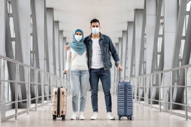 Havalimanında bavulla poz veren mutlu Müslüman çift portresi, Terminalde duran İslami eşler, Salgın ve Serbest Alan sırasında birlikte seyahat eden