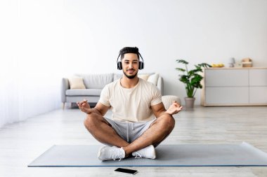 Kulaklığı ve cep telefonu olan genç bir Arap evde nilüfer yogası pozu verirken müzik dinliyor. Modern aletler kullanan, kapalı alanda çalışan, uzayı kopyalayan tam uzunluktaki doğulu adam.