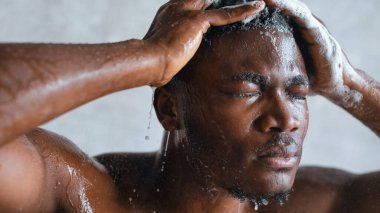 Bir adam saçını yıkarken banyoda duruyor. Başının üstündeki duş başlığından su akıyor. Ellerini başının üstünde tutuyor, gözleri kapalı, saçlarında sabunla..
