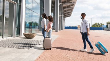 Coronavirus Seyahatleri. Koruyucu tıbbi maskeli siyahi yolcular havaalanı binasında bavullarıyla yürürken, covid salgını nedeniyle sosyal mesafelere göre giriş kuyruğunda bekliyorlar. Yeni Normal Kurallar