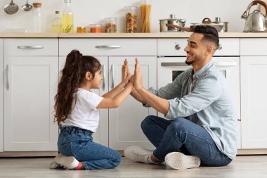 Güzel bir ortadoğu aile babası ve kızı mutfakta oynuyor, yerde oturuyor ve el çırpıyor. Neşeli Arap baba ve küçük kız çocuk evde, yan görüşte, kopyalama alanında birlikte eğleniyorlar.