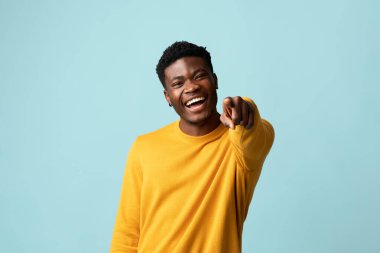 Mavi stüdyo arka planında poz verip kameraya gülüp el kol hareketi yapan, Afro-Amerikan genç adam işaret parmağı gösteriyor, flört ediyor, mekanı kopyalıyor.
