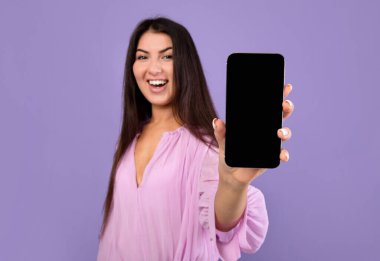 Mobil uygulama kavramı. Heyecanlı Ermeni kadın mor arka planda boş ekranlı akıllı telefon gösteriyor, seçici odaklanma. Gülümseyen ve cep telefonunu boş ekranla tutan bayanın yakın çekimi