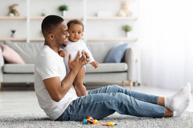 Bebekle bağ kurmak. Mutlu genç siyah adam evde küçük çocuğuyla oynuyor, Afrikalı Amerikalı babayı seviyor, sevimli küçük çocukla vakit geçiriyor, oturma odasında yerde dinleniyor.