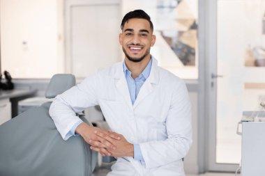 Yakışıklı bir Arap stomatolojistin portresi, diş hekiminde poz veren, üniformalı orta doğulu dişçi, iş yerinde durup kameraya bakıyor, hastayı kontrol etmeye hazır.